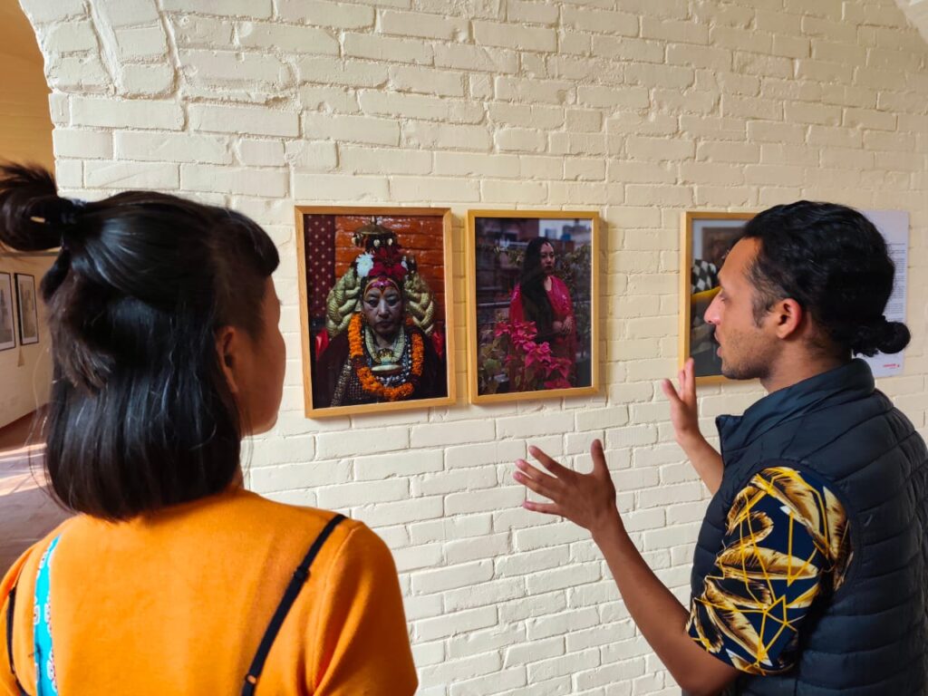 A man is looking at two photographs hanging on the wall and is describing it to a woman standing next to him. His hands are spread out as he provides context to the work as the woman intently listens.
